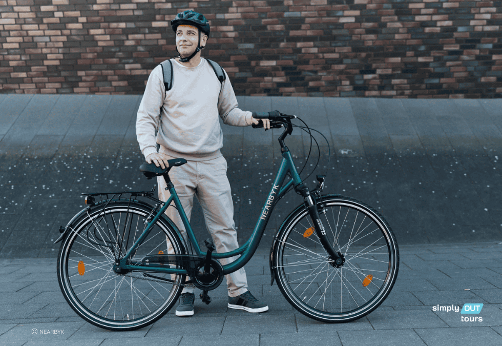 Mann mit Helm und Rucksack steht lächelnd neben einem dunkelgrünen Citybike von Nearbyk auf dem Gelände der Zeche Zollverein; rechts moderne Ziegelwand, links farblich abgestimmter Hintergrund in grün – Teil der neuen Leihrad-Flotte von simply out tours.